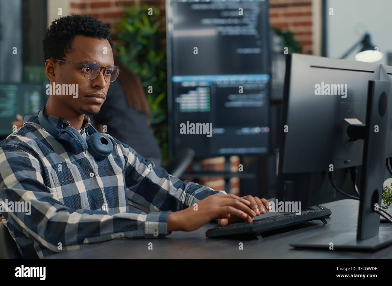 Focused programmer fixes his glasses while writing code at a desk, with teammates programming user interface work in the background. Stock Photo