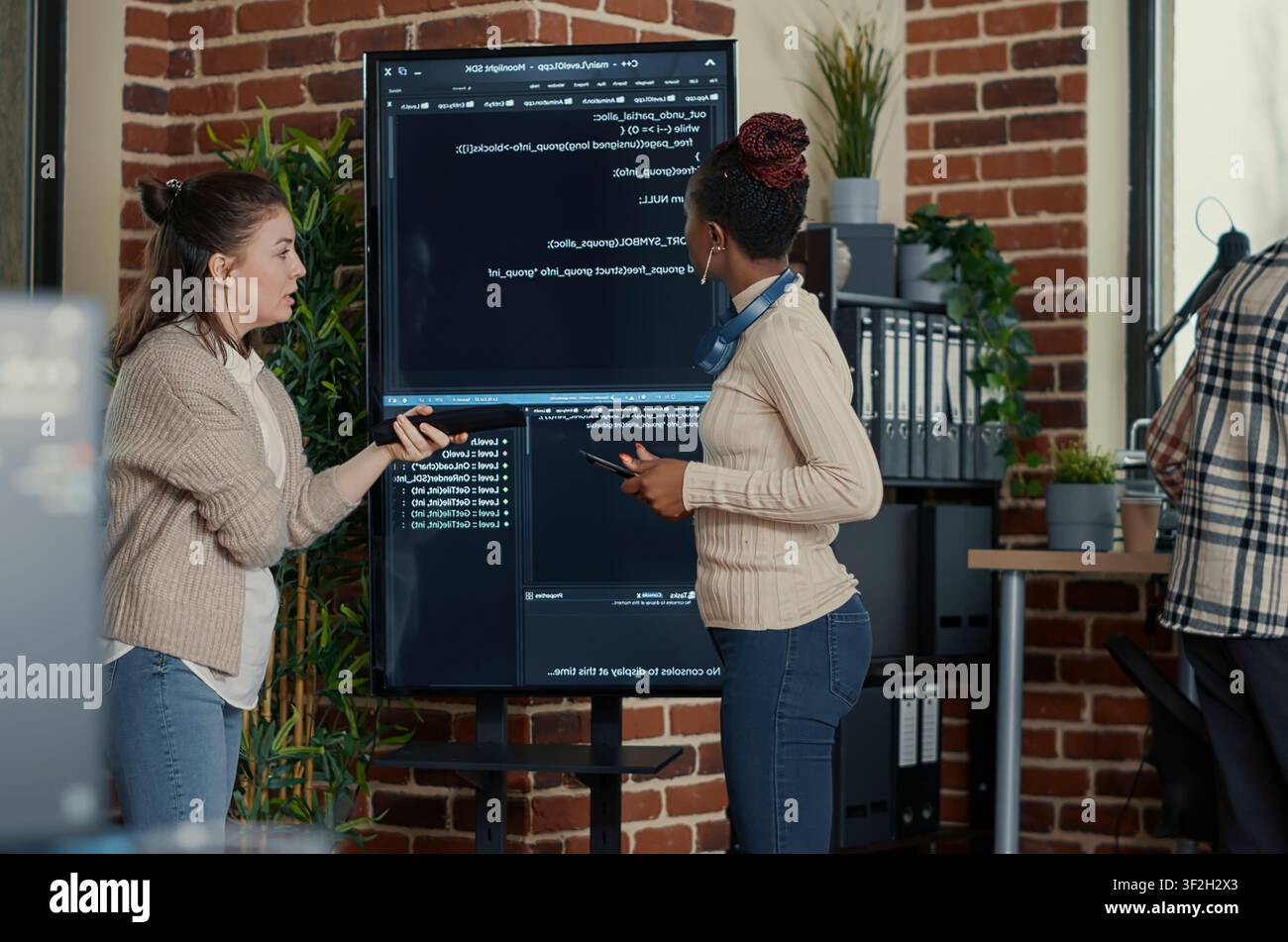 Programmer holding digital tablet analyzing code on wall screen tv while colleague is looking for errors pointing using remote control, with wall screen and African American. Stock Photo