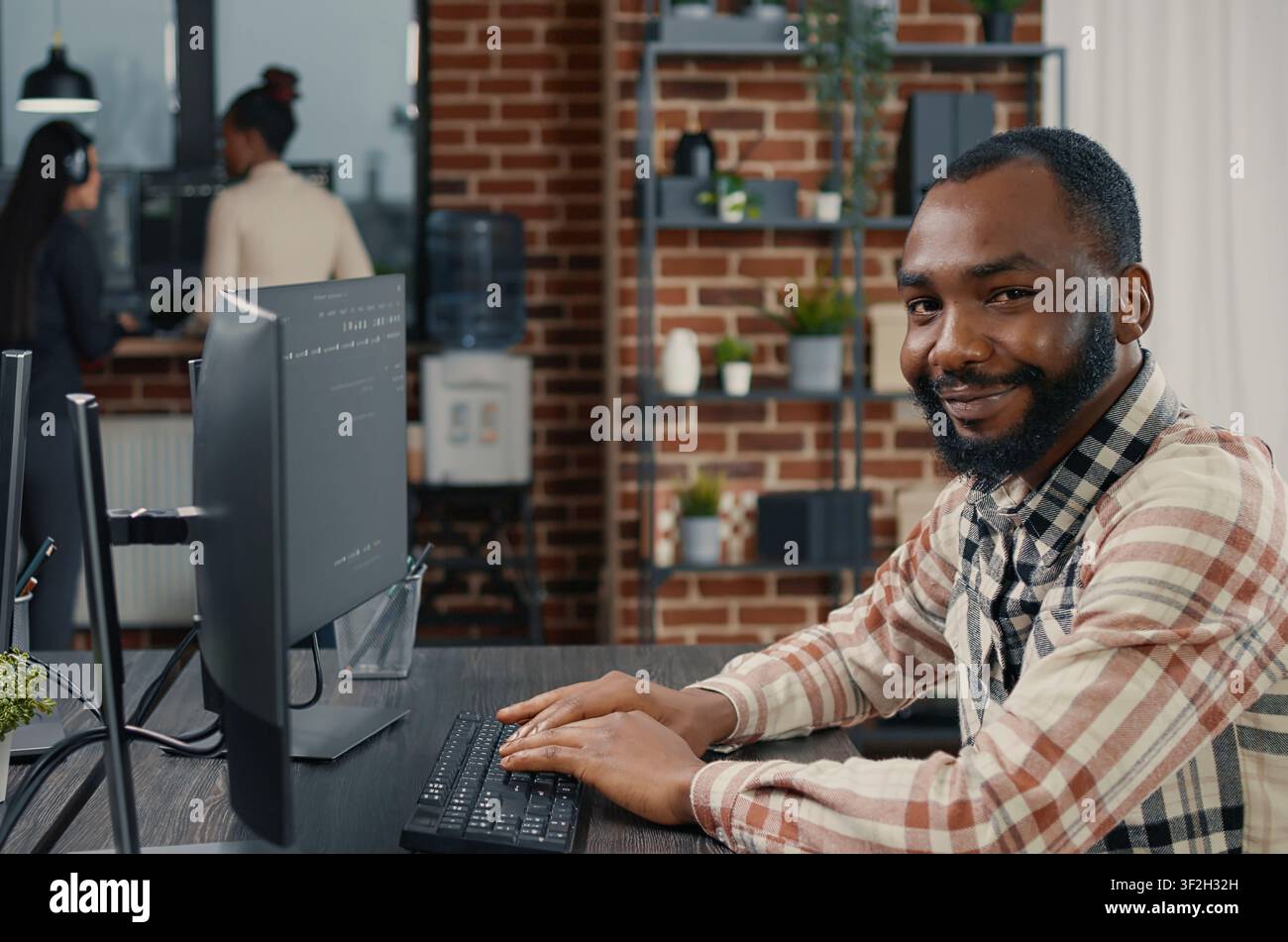 Database programmer writing code and smiling while coworker walks by holding clipboard in it agency, relaxed app developer typing on keyboard with colleagues computing big data. Stock Photo