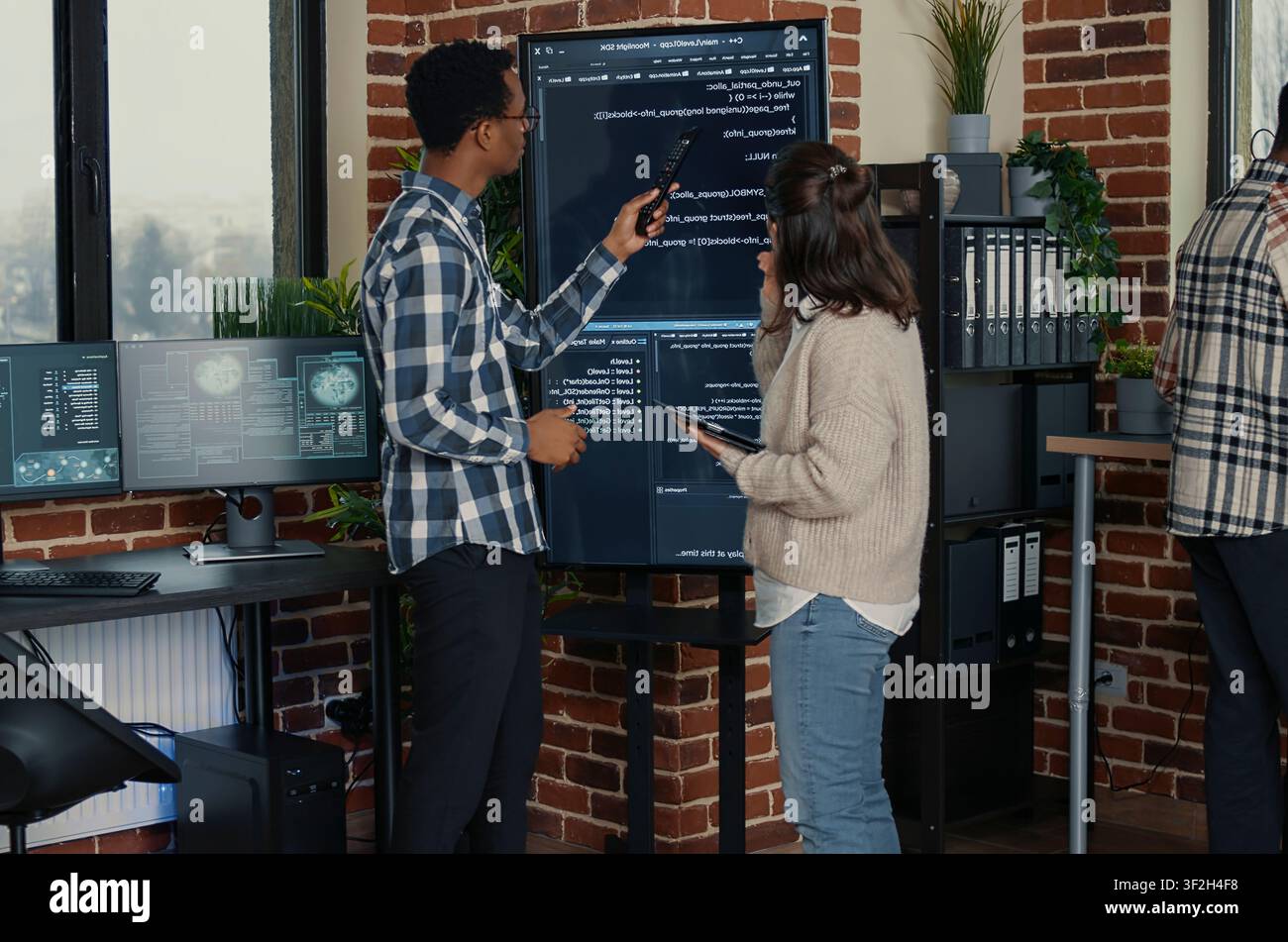 Database administrators compare source code on a wall screen and tablet in a busy server room while cloud programmers debug software algorithms. Stock Photo