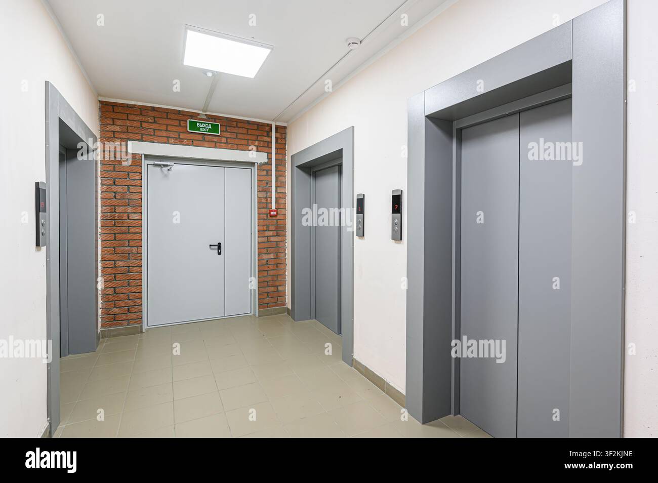An interior hallway with two elevators, an exit door framed by brick, and tile flooring Stock Photo