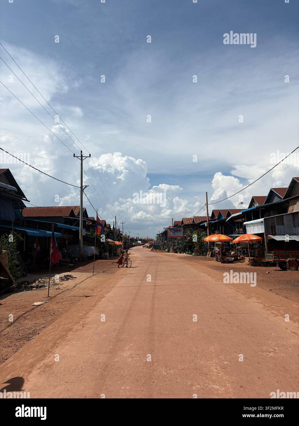 18 April 2025, Prasat Bakong, Siem Reap, Cambodia - Dusty dirt road lined with local shops and utility poles near Bakong temple village under dramatic cumulus clouds. - Smartphone Captured Stock Image