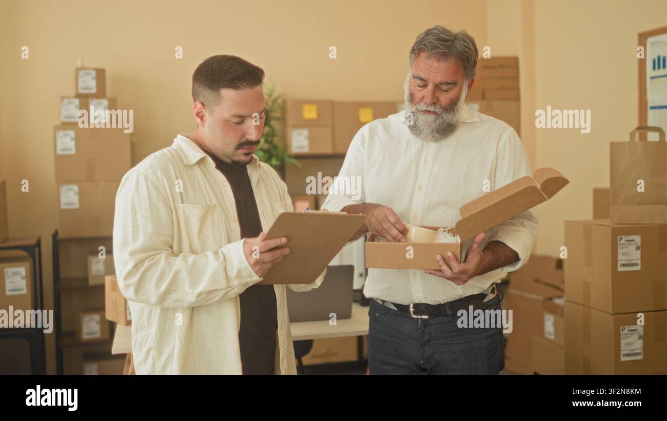 Two men inspect and hold a parcel box while one reads a clipboard in a small business building; teamwork collaboration efficiency. Stock Photo