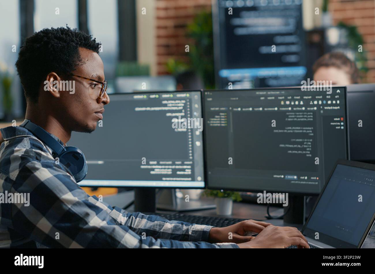 Software developer thinking while touching beard while typing on laptop sitting at desk with multiple screens parsing code, focused database admin working with team coding in the background. Stock Photo