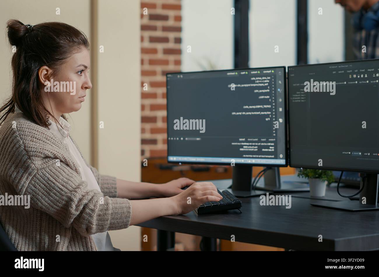 Focused programmer writes code while looking at multiple computer screens, with programming language algorithms and teamwork in the background. Stock Photo