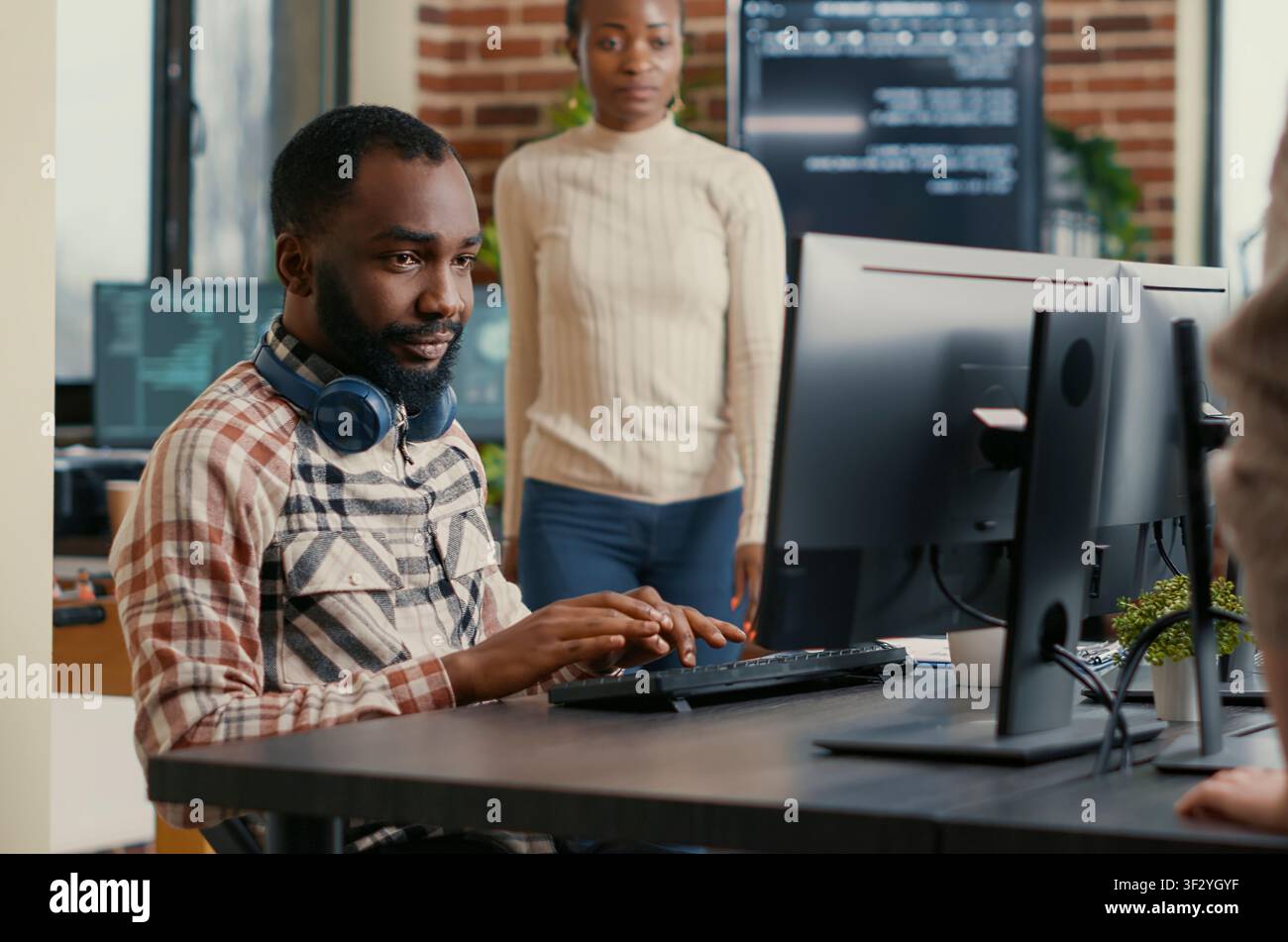 Smiling software developer wearing wireless headphones smiling working at computer screen while typing at a computer, showing a smiling developer workflow with code, headphones and IT office screens. Stock Photo