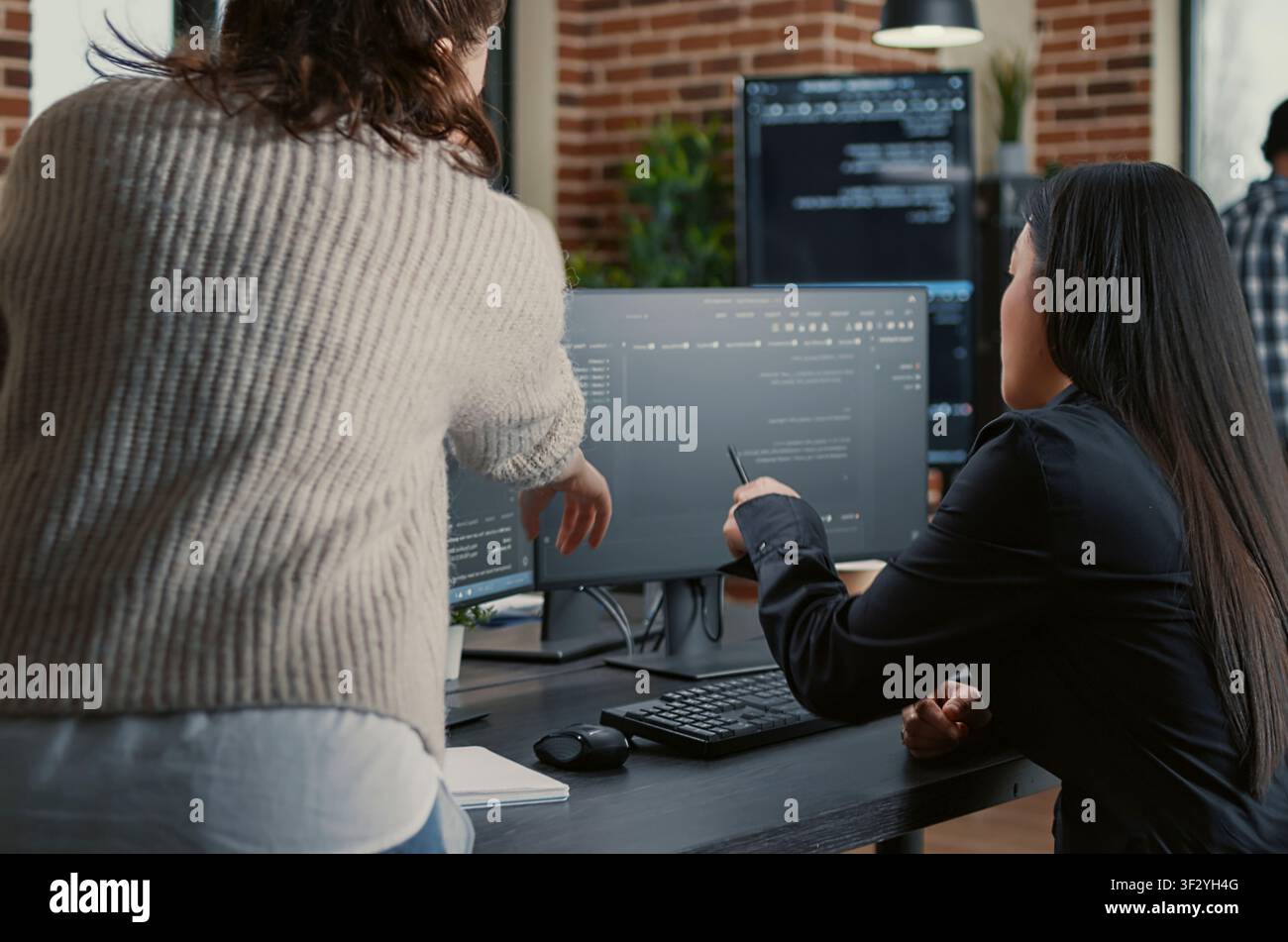 Programmer pointing pencil at computer screen with software compiling code explaining source algorithm to coworker standing next to desk around computer screens, showing developer teamwork, source. Stock Photo