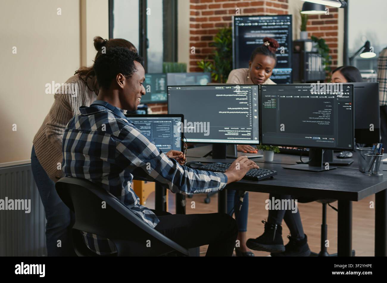 Software developer reviews running code on multiple screens, takes off his glasses and celebrates with a colleague high five during a successful coding moment. Stock Photo