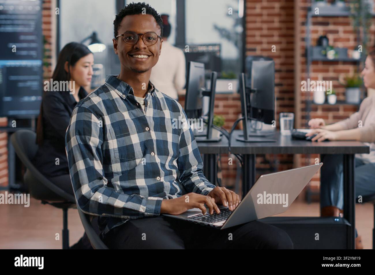Portrait of artificial intelligence app developer sitting down typing on laptop fixing glasses looking up and smiling at camera, programer using portable computer innovating cloud computing. Stock Photo