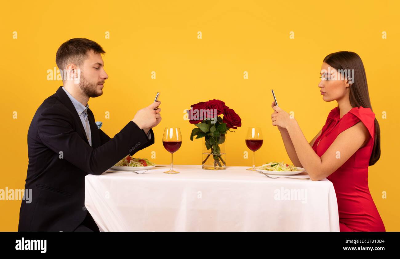 Couple Using Cellphones Sitting At Table Posing Over Yellow Background Stock Photo