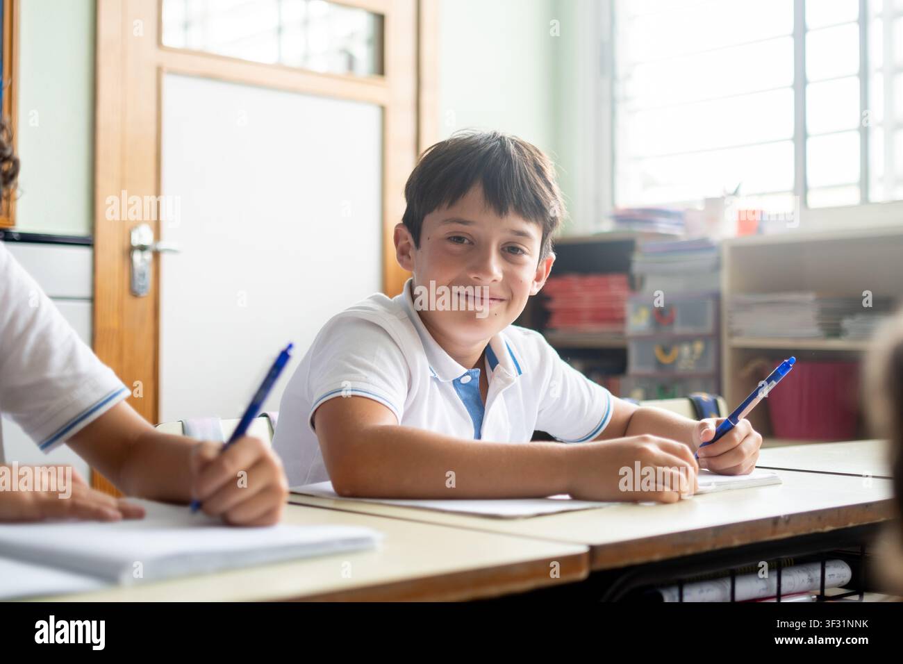 Smiling elementary school boy looking at the camera while writing in his notebook during class. Stock Photo