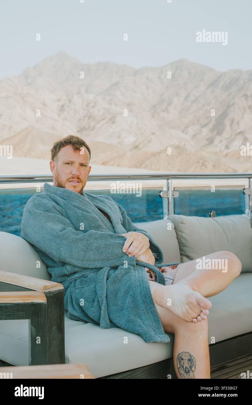 Man in bathrobe sitting on yacht deck with mountains in background Stock Photo