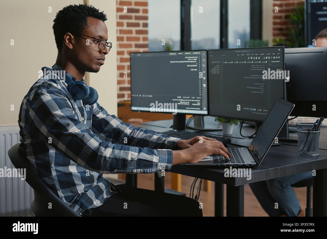 Programmer thinking while touching beard and fixing glasses while typing on laptop sitting at desk with multiple screens parsing code around computer screens, showing developer teamwork, source. Stock Photo