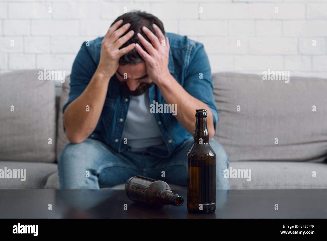 Sad man holds his head and sitting on couch, empty bottles on table, close up Stock Photo