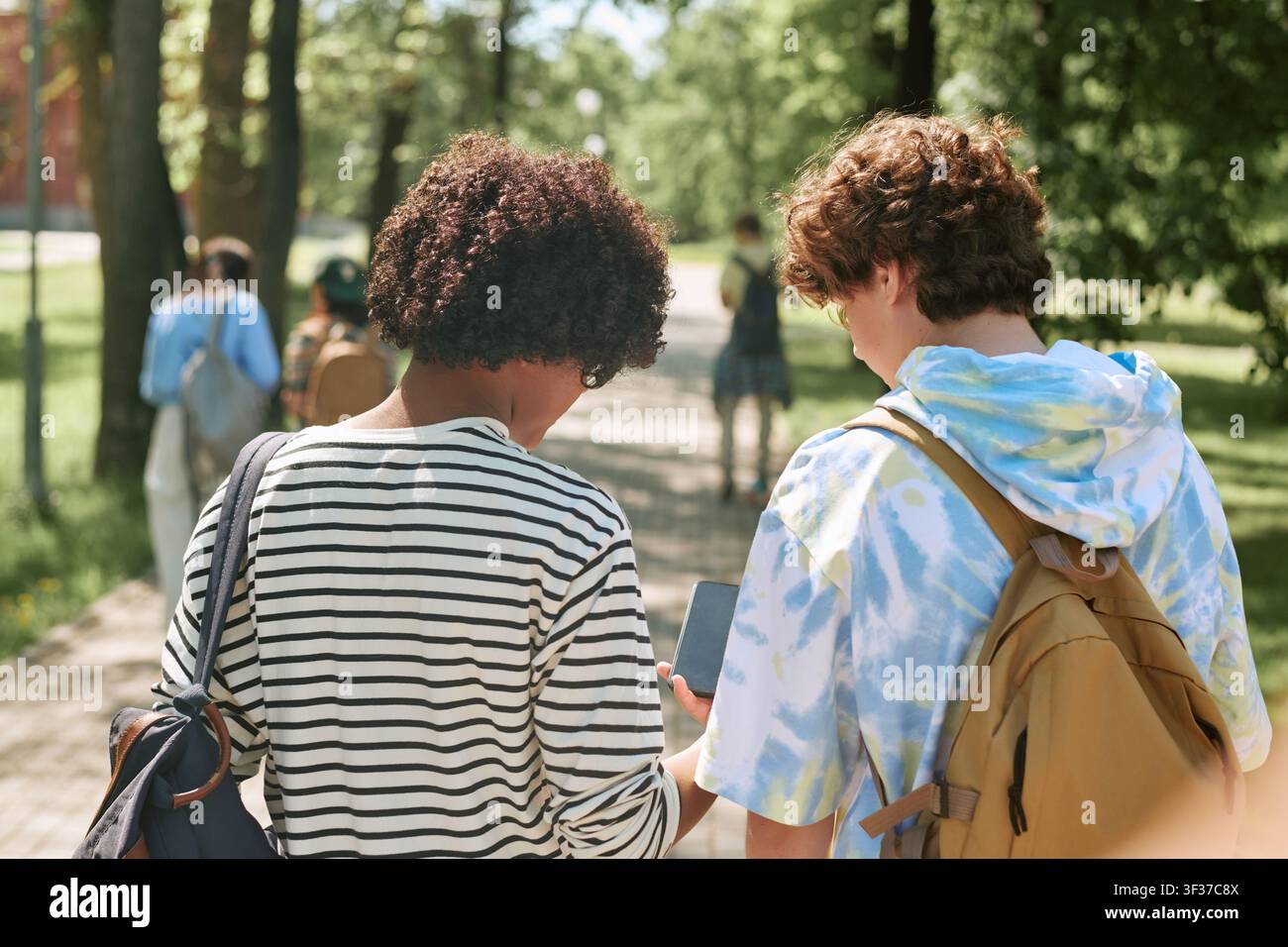 Diverse Teen Students Using Smartphone while Walking on Campus Path Stock Photo