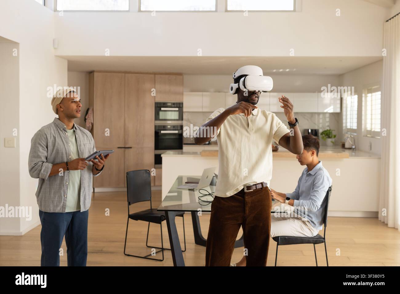 Diverse male coworkers testing VR headset, watching tablet, typing on laptop in kitchen Stock Photo