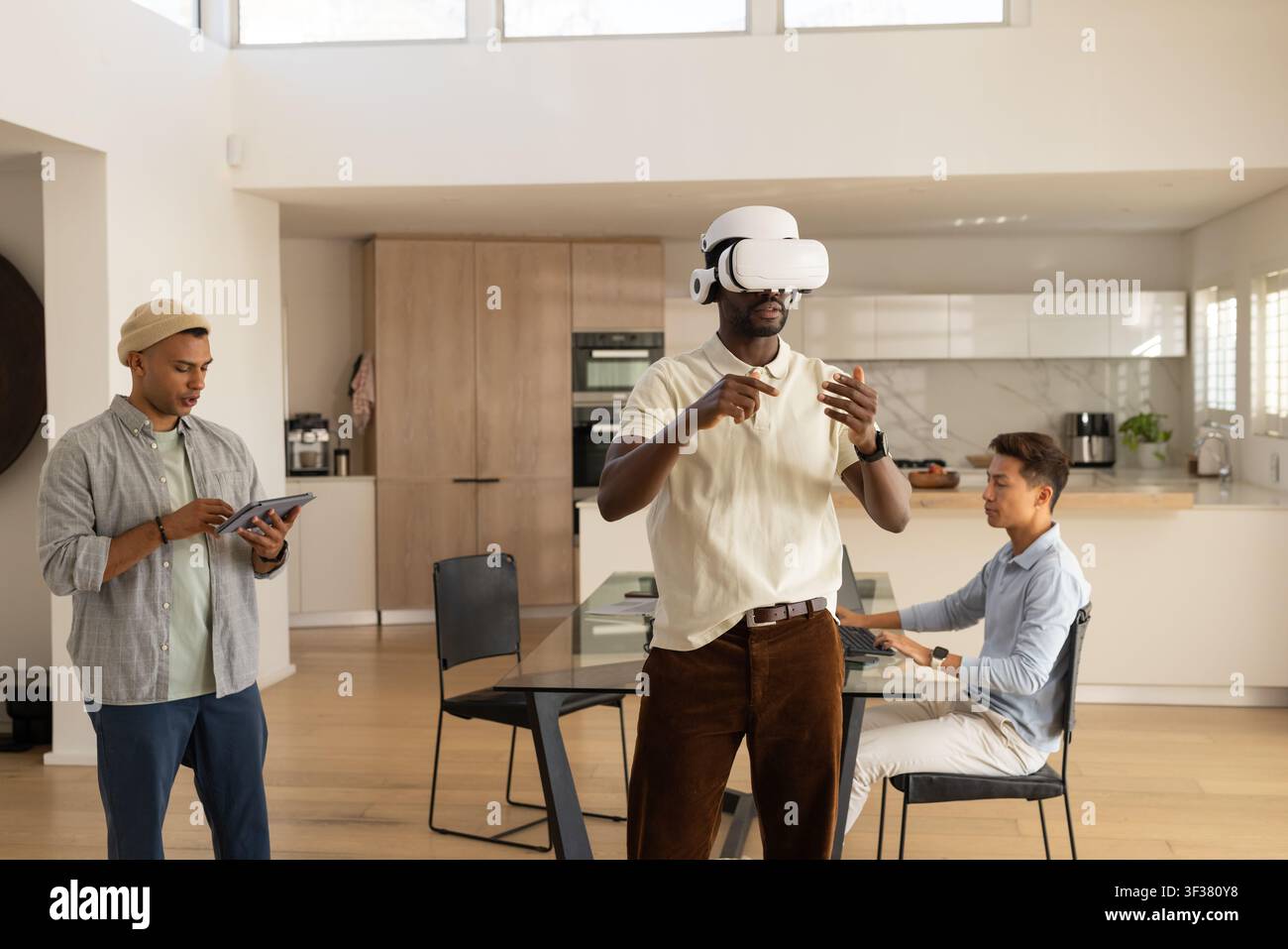 Diverse male coworkers testing white VR headset while using tablet and laptop in kitchen Stock Photo