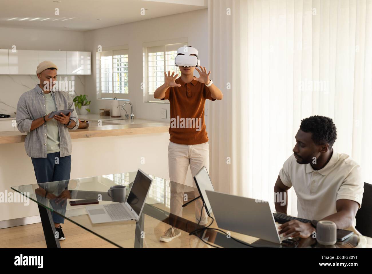 Diverse male coworkers in kitchen using laptops tablet Asian man wearing white VR headset reaching Stock Photo