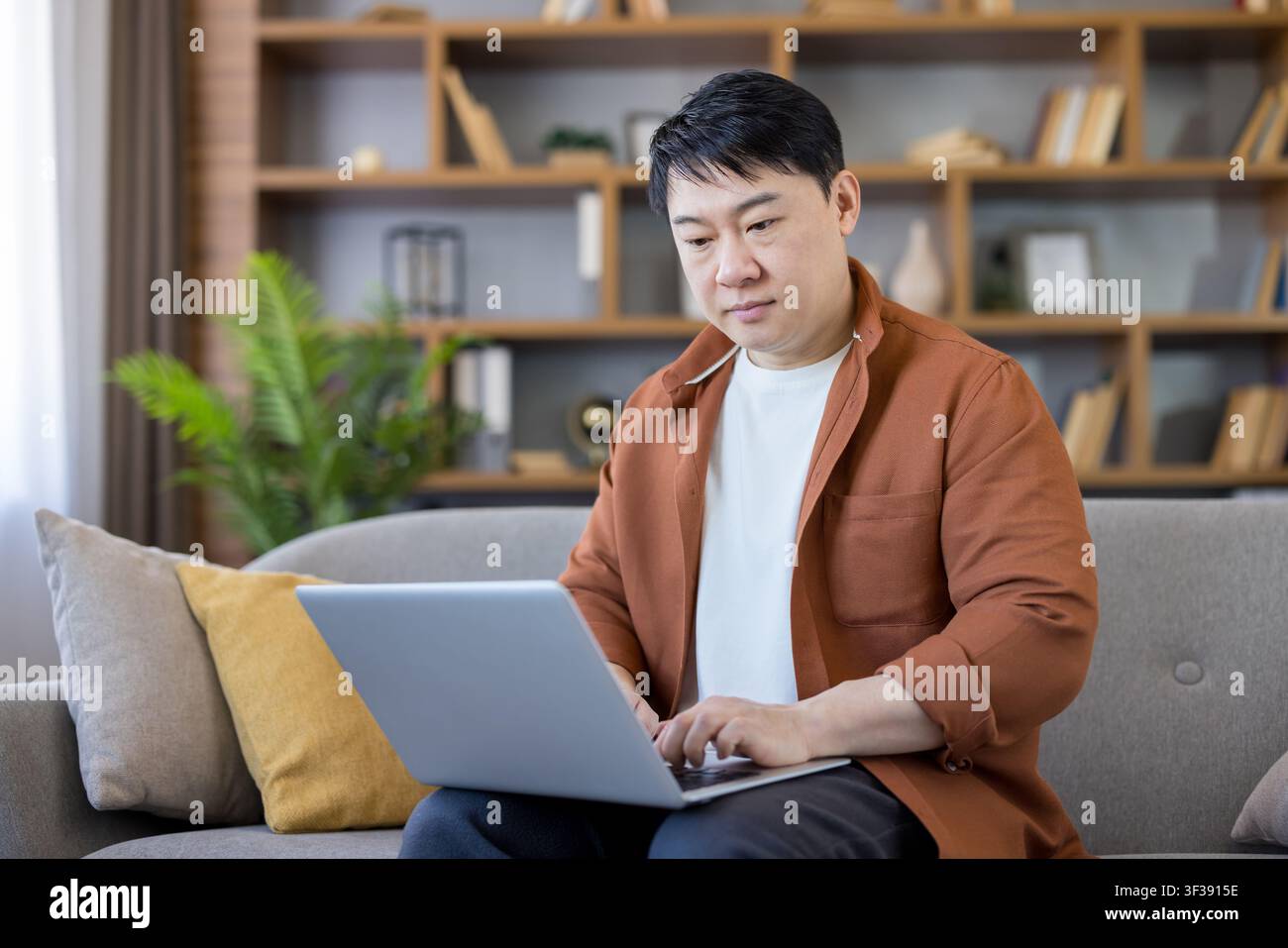 Asian man sitting comfortably on sofa, typing on laptop, engaging in remote work, online learning, or casual browsing at home Stock Photo