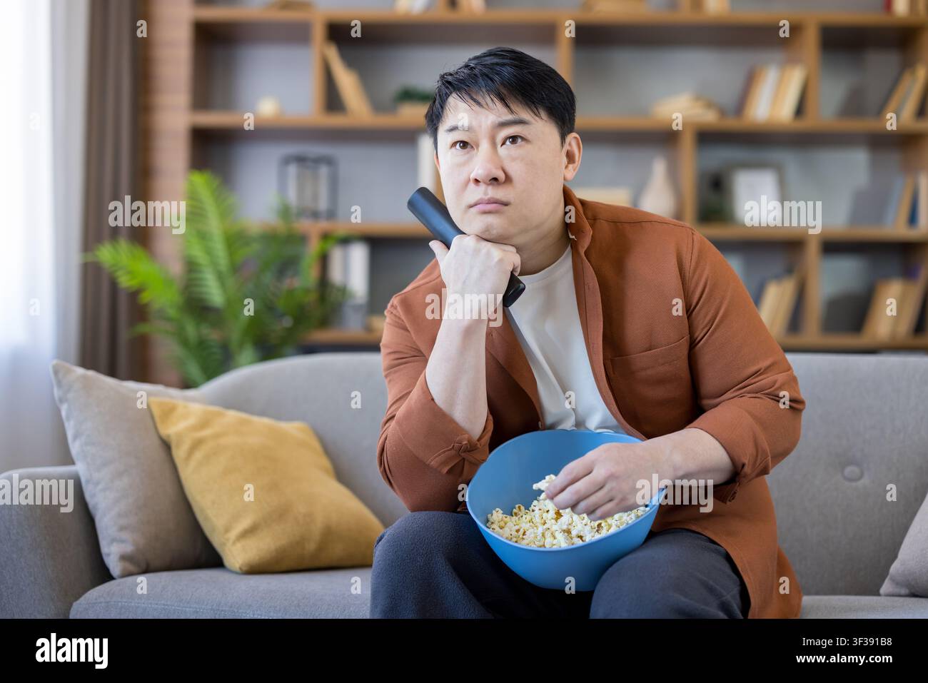 Asian man feeling bored and unimpressed while sitting on a comfortable couch in his living room, holding a remote control and eating fresh popcorn, relaxing at home during leisure time Stock Photo