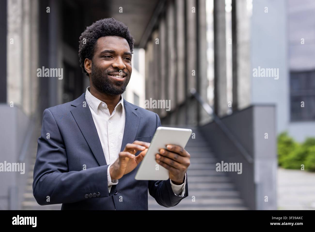 Young businessman standing confidently outside a modern office building, smiling while using a digital tablet, representing success, technology, and professional communication Stock Photo