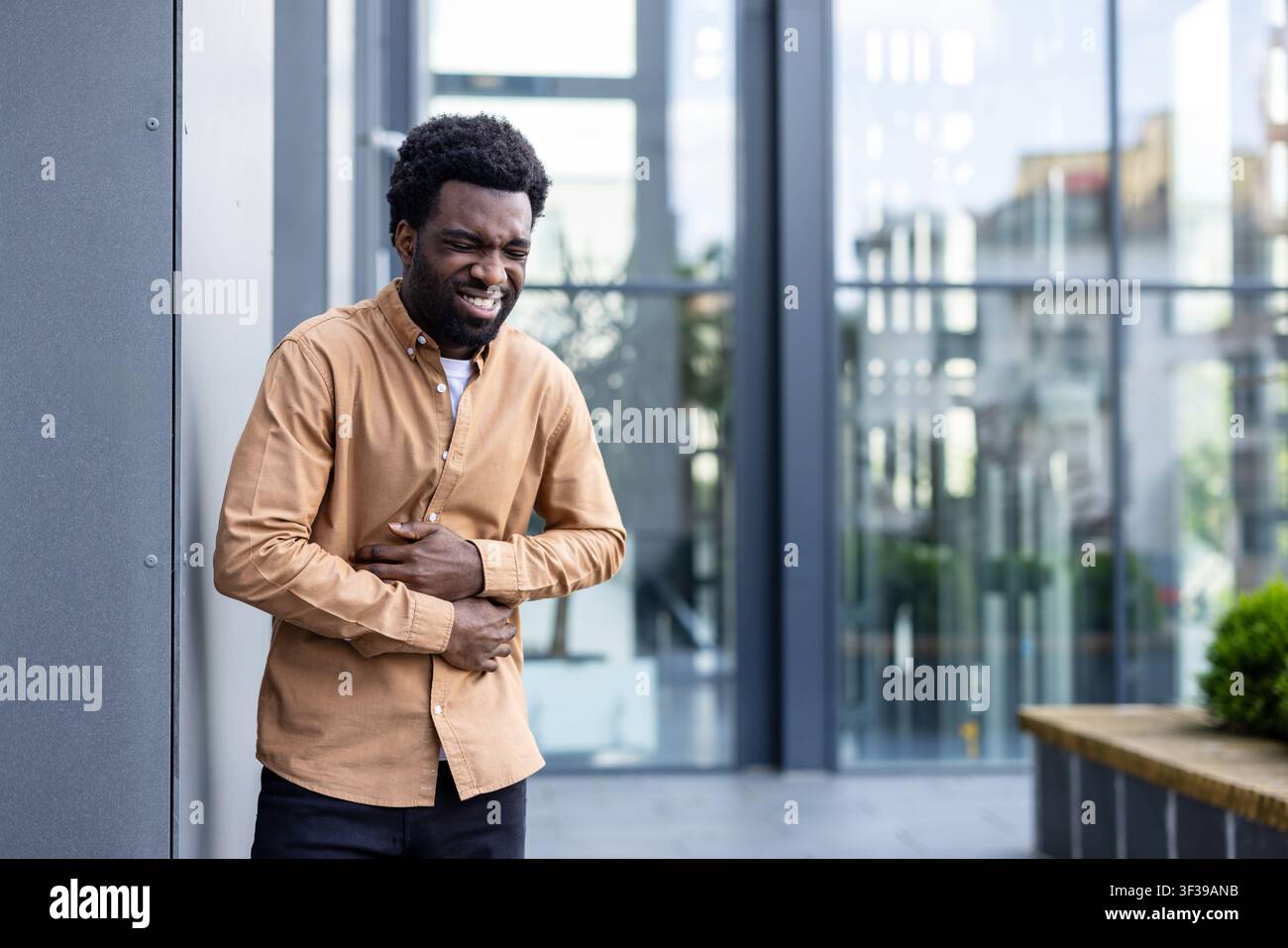 Young man standing outdoors, expressing discomfort and pain, holding his stomach with both hands while suffering from indigestion or cramps near an office building Stock Photo