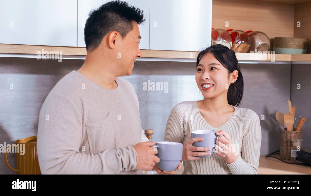Happy Asian Couple Smiling And Talking While Holding Coffee Mugs In Kitchen, Home Concept Stock Photo