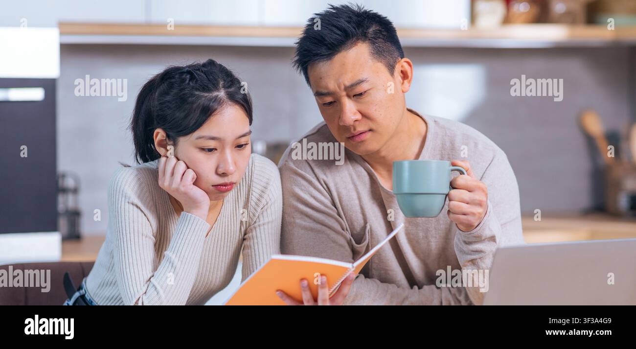 Focused Asian Couple Reviewing Documents In Modern Kitchen, Serious Discussion Stock Photo