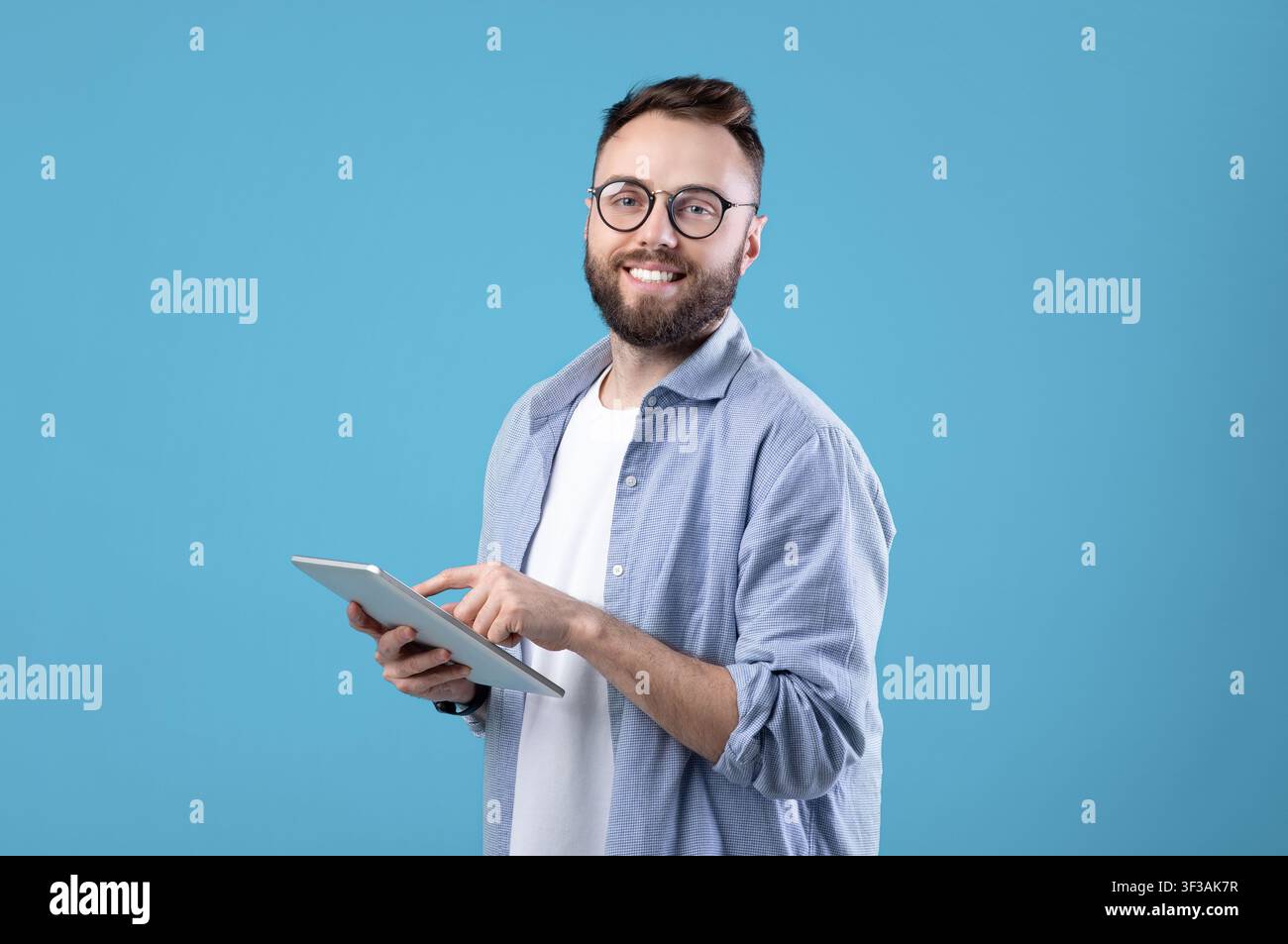Positive young guy in casual wear using tablet computer over blue studio background Stock Photo