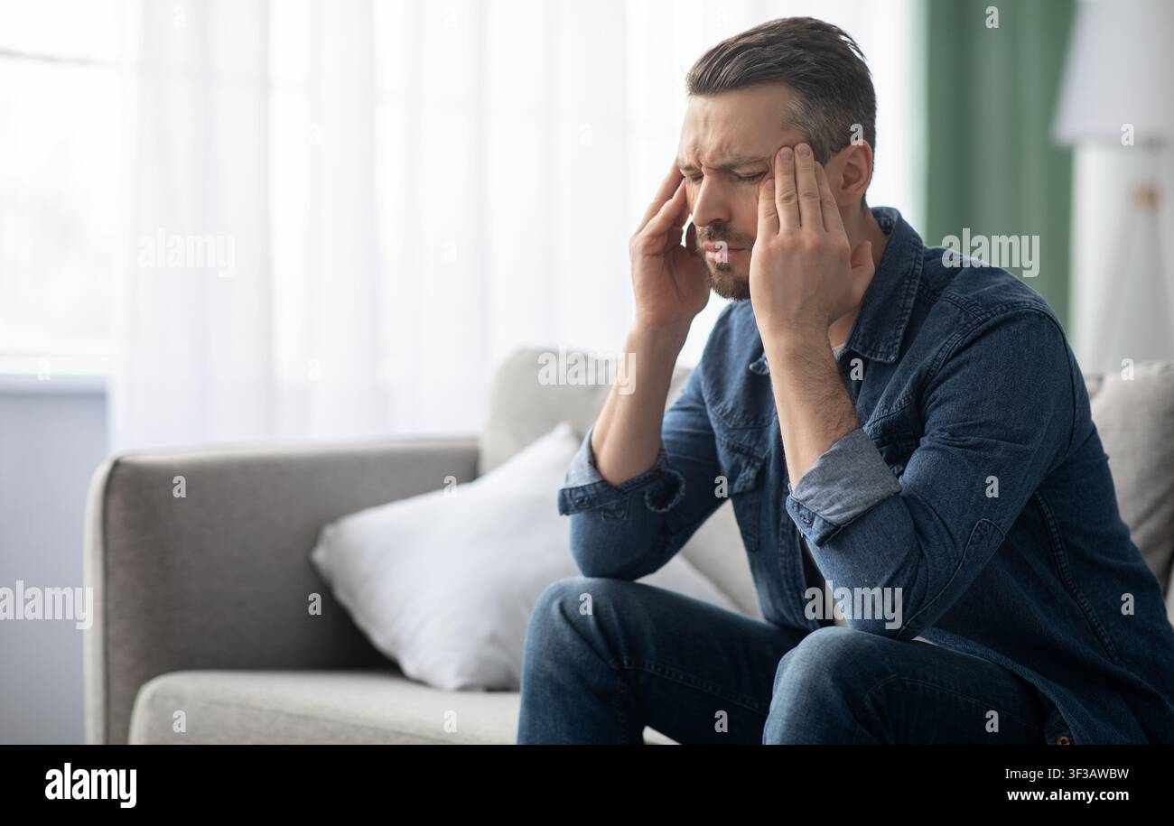 Stressed man having headache, sitting on sofa at home Stock Photo