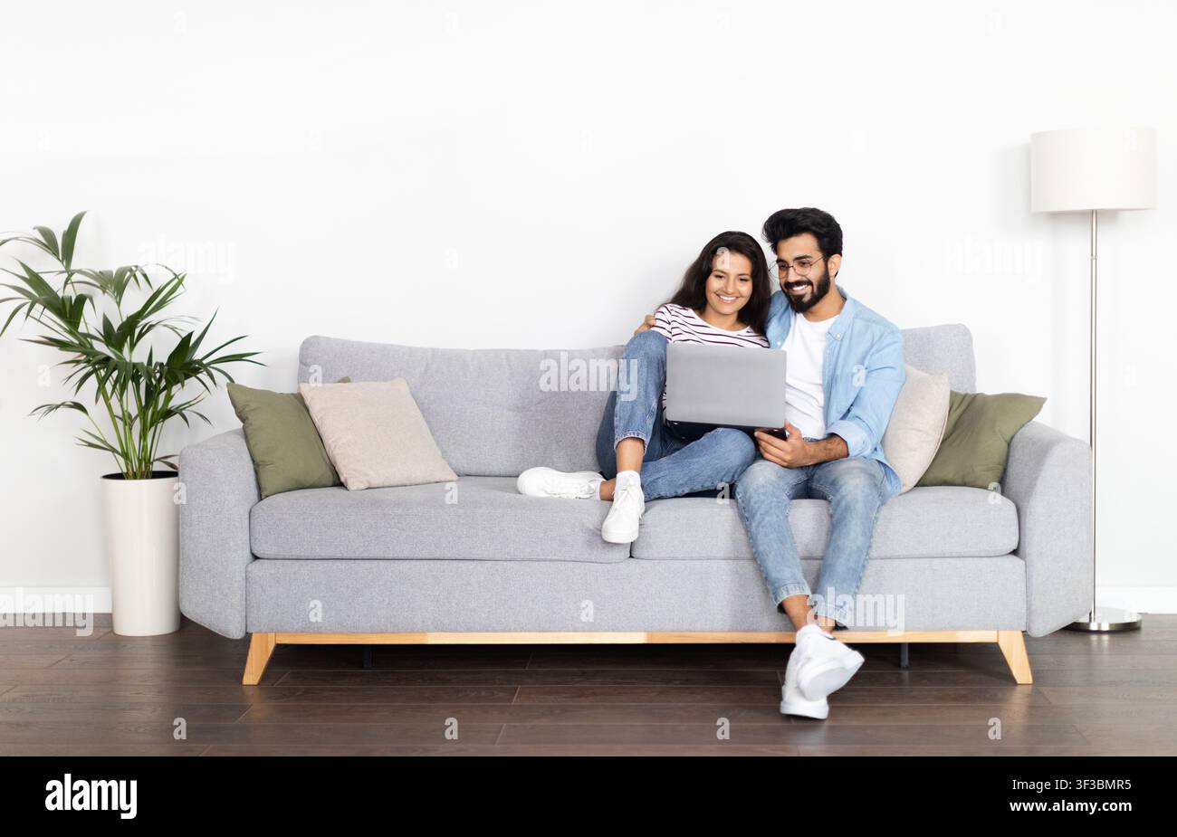 Loving eastern couple sitting on couch at home, using laptop Stock Photo