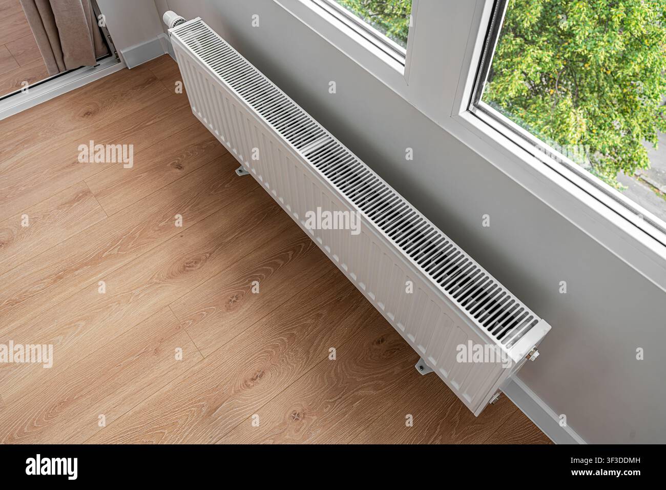 A modern heating radiator is mounted under a window in a bright room with light wooden flooring. Stock Photo