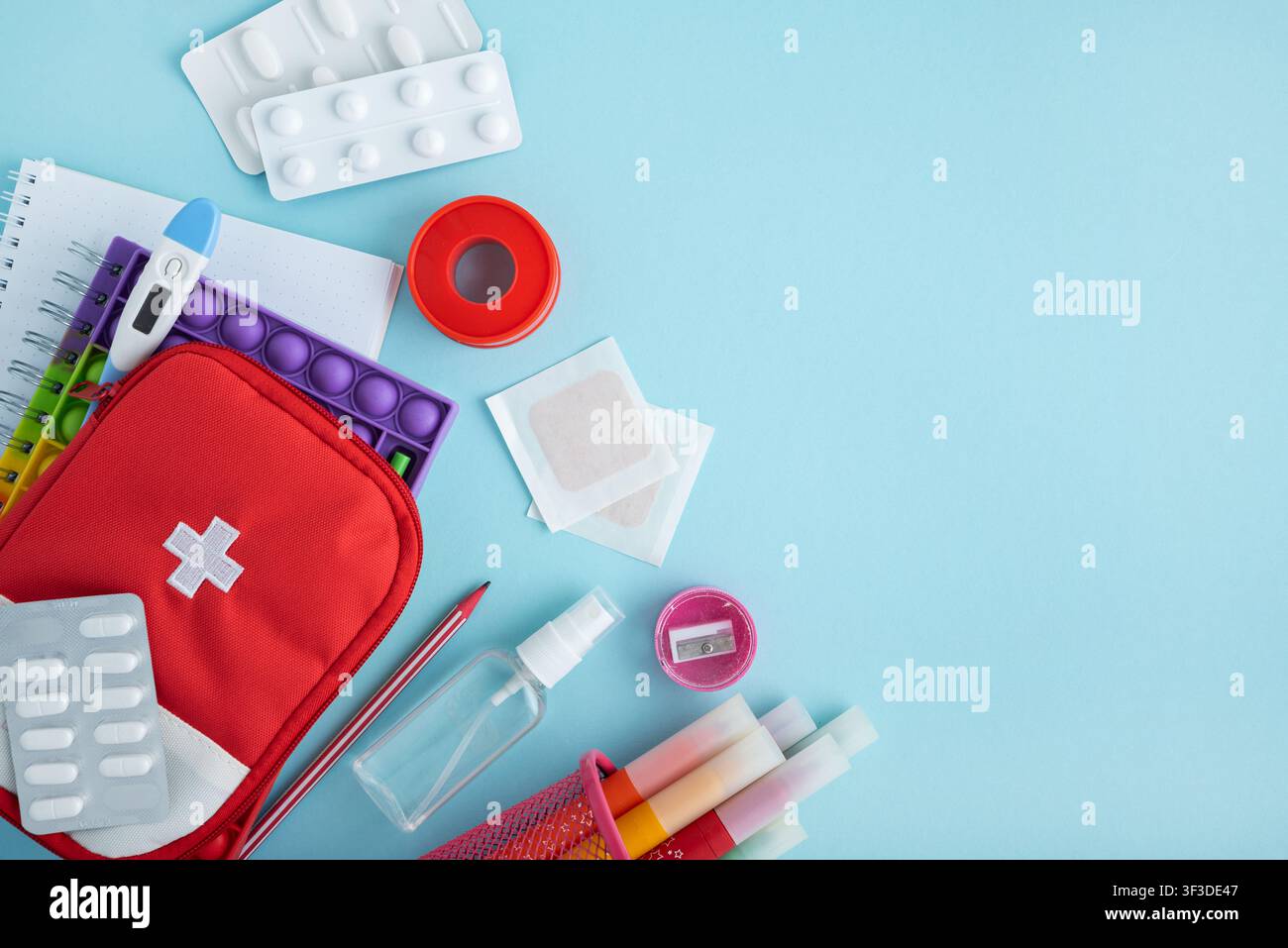 Red first aid kit bag with cross symbol, various pills, thermometer, adhesive bandages, and colorful school learning supplies on a light blue backgrou Stock Photo