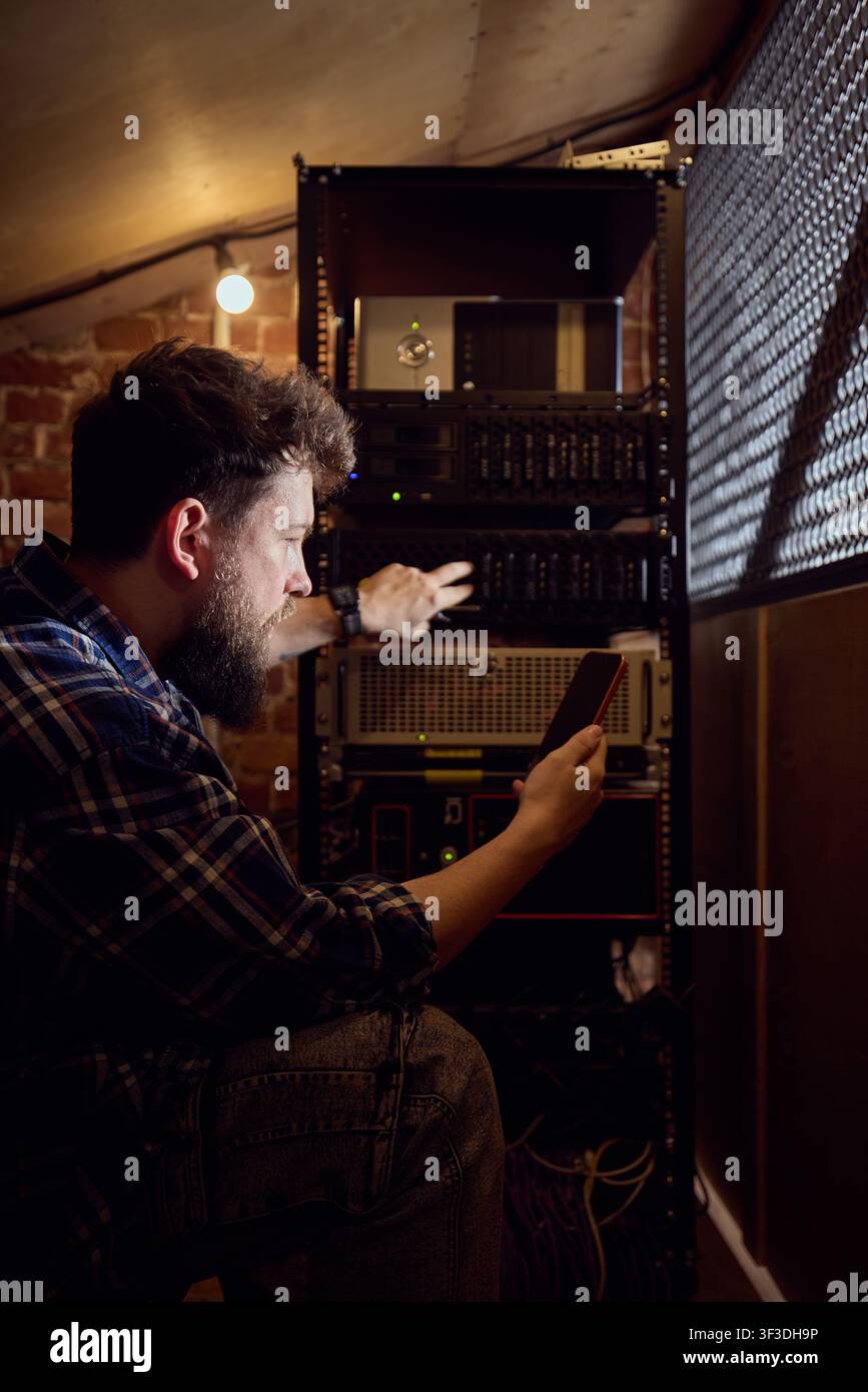 Male IT technician checking server rack with smartphone in dim technical room Stock Photo