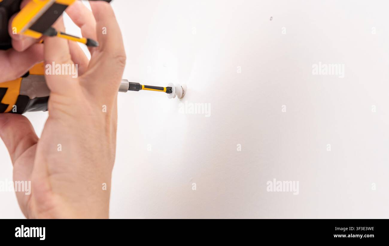 Mans hand using yellow cordless drill to drive screw into white wall for bracket installation. Home improvement, mounting shelf support, DIY fastening Stock Photo