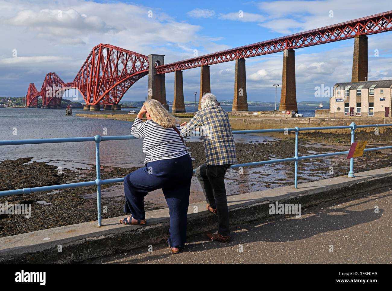 Visitors admire the red cantilever Forth Bridge from South Queensferry promenade on a bright summer day beside the Firth of Forth. EH30 9HN Stock Photo