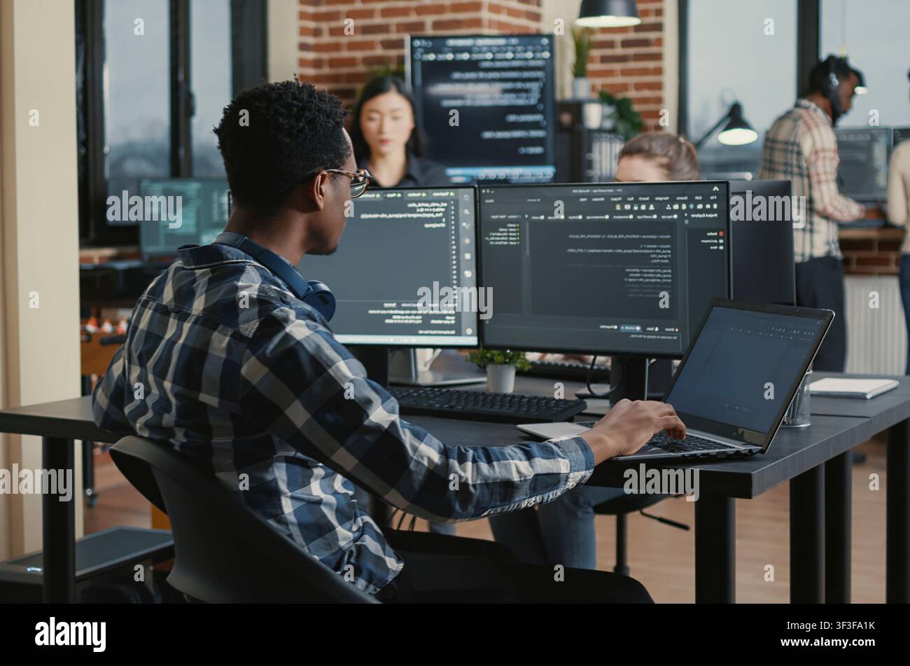 Programmer fixes his glasses while typing on a laptop and checking compiled code results at a desk with multiple software screens. Stock Photo