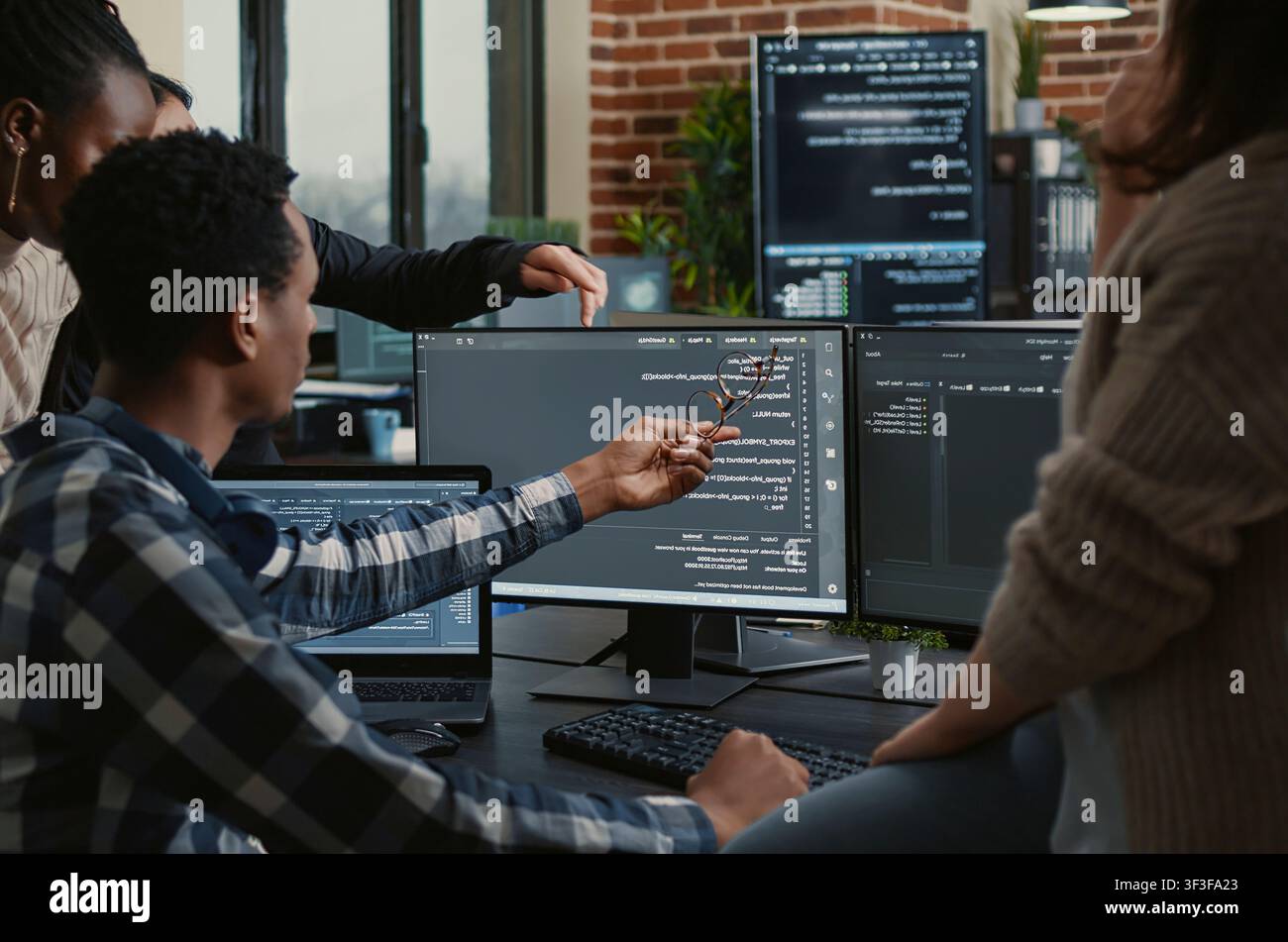 Software developers review source code compiling errors with the team, asking for explanations in front of multiple screens running algorithms. Stock Photo