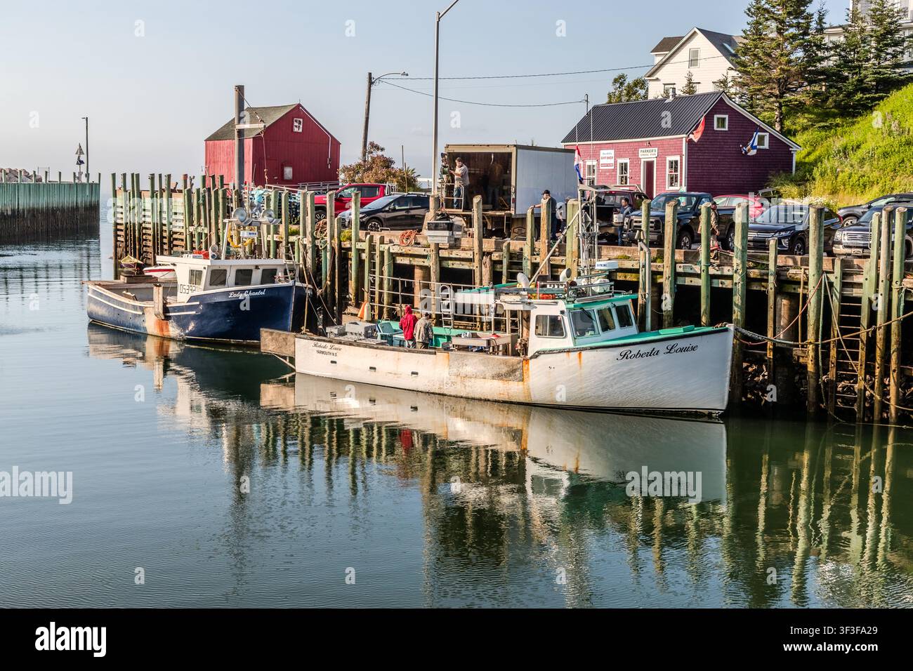 Fishing boats, including the cutter ‘Roberta Louise’, are moored at the quay in Halls Harbour, Nova Scotia, Canada, in front of the red-painted boathouses and storage buildings of the fishing village. Here, the Bay of Fundy has a tidal range of up to 15 metres between low and high tide Stock Photo