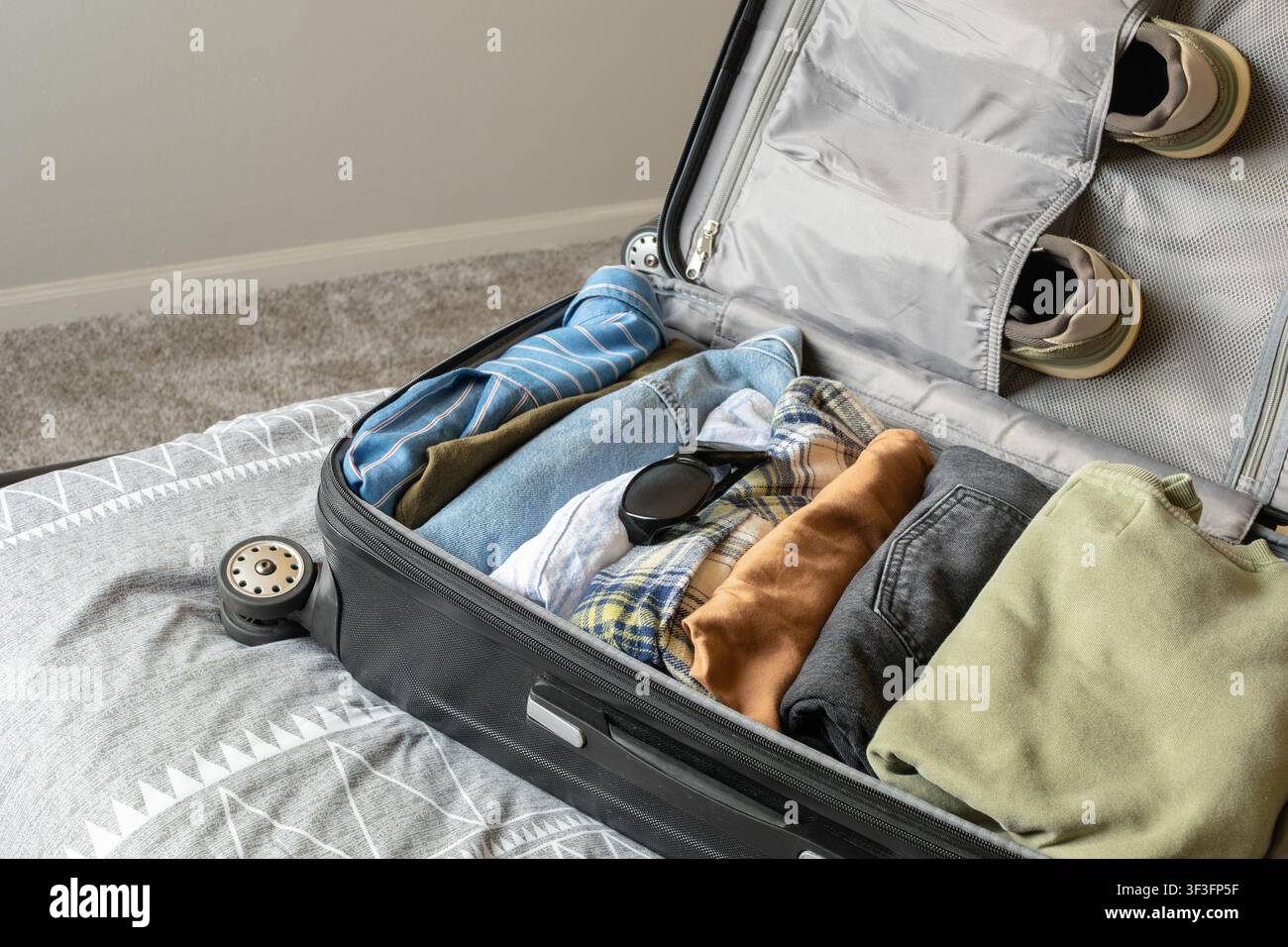 An open black suitcase lying on a grey bedsheet, carefully packed for a trip. Inside the luggage are shirts, jeans, blue denim jacket, sunglasses and Stock Photo