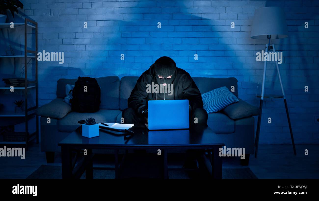Masked hacker downloading private information using laptop Stock Photo