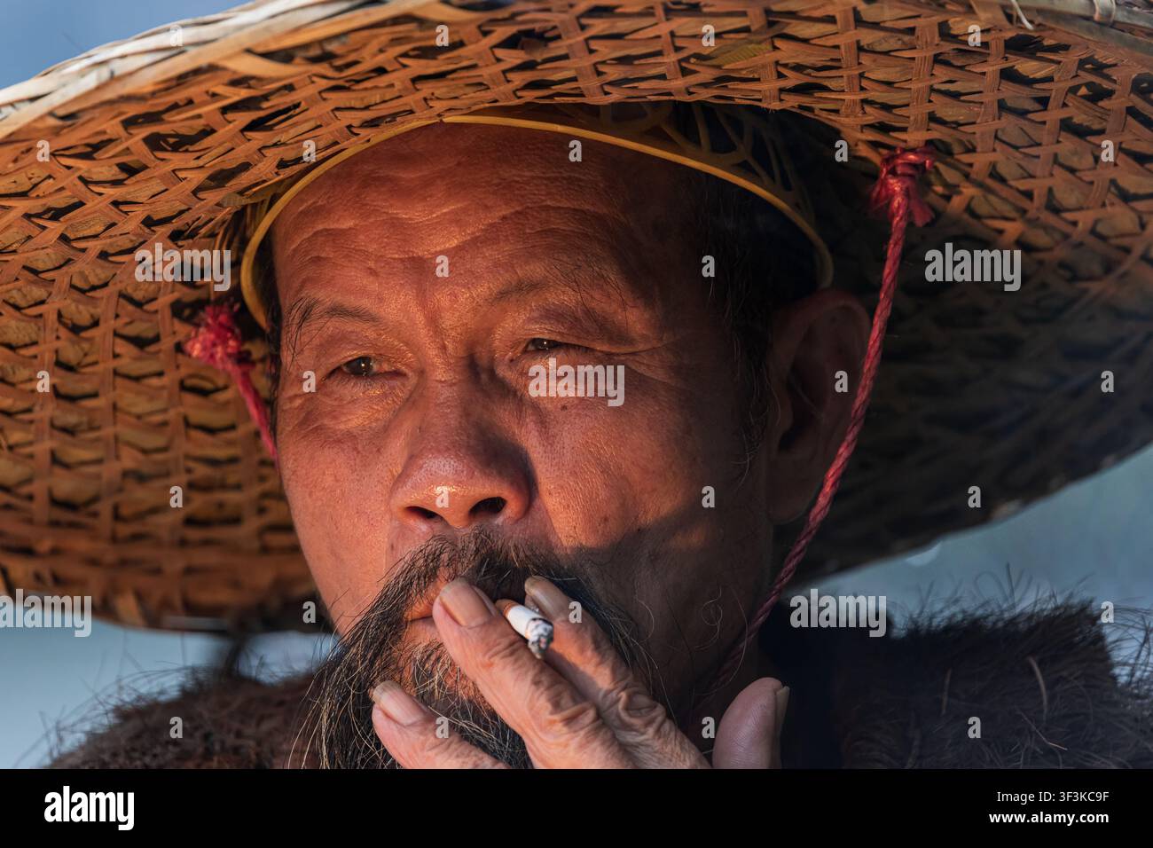 Xingping, Guangxi, China - 18 June 2026: View of an elderly man wearing a traditional woven straw hat while smoking a cigarette in Xingping Ancient To Stock Photo