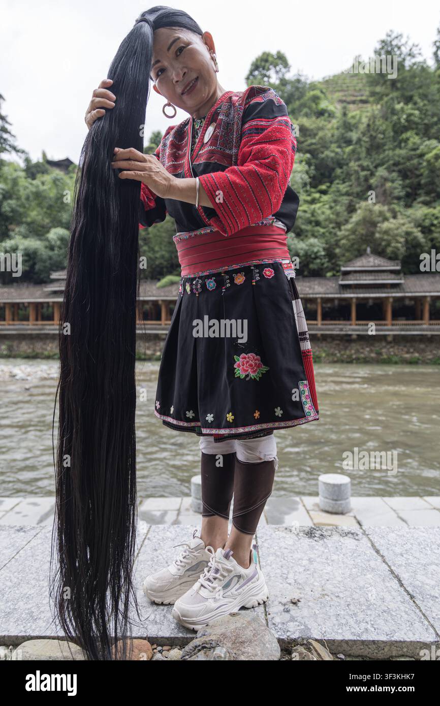 Guangxi, China - 18 June 2026: View of a Red Yao woman in traditional embroidered clothing holding her exceptionally long black hair, standing beside Stock Photo