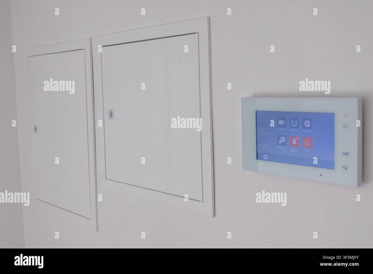 A sleek, white intercom panel with a blue touchscreen interface is installed on a clean, minimalist wall next to access panels Stock Photo