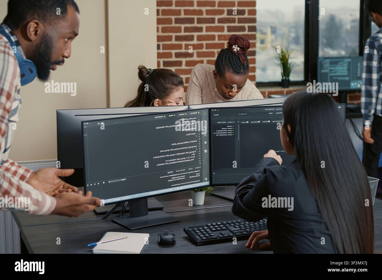 Mixed programmer team works on a group software project across multiple screens, with a coder pointing at code while discussing development tasks in an IT office. Stock Photo