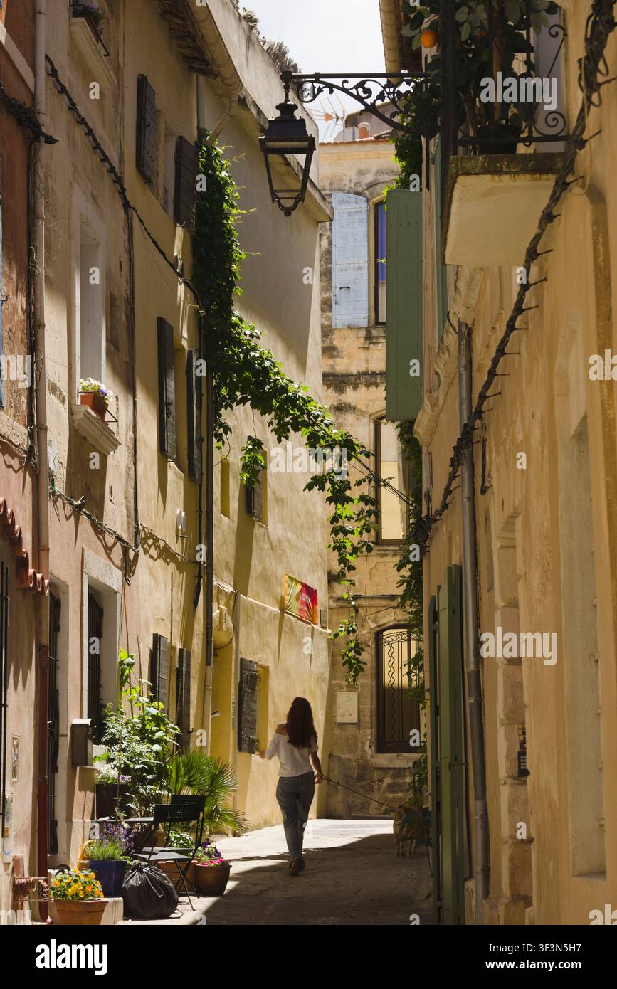 A woman walks her dog down a narrow, sunlit historic alley lined with traditional Provencal buildings in Arles, France. Stock Photo