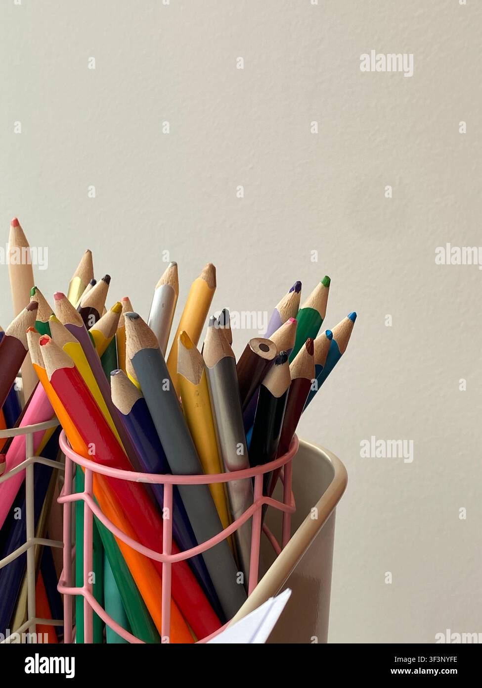 Bright colored pencils lean in a pink wire holder. Soft light highlights every tip and hue. Simple background emphasizes creativity and choice. Ideal Stock Photo