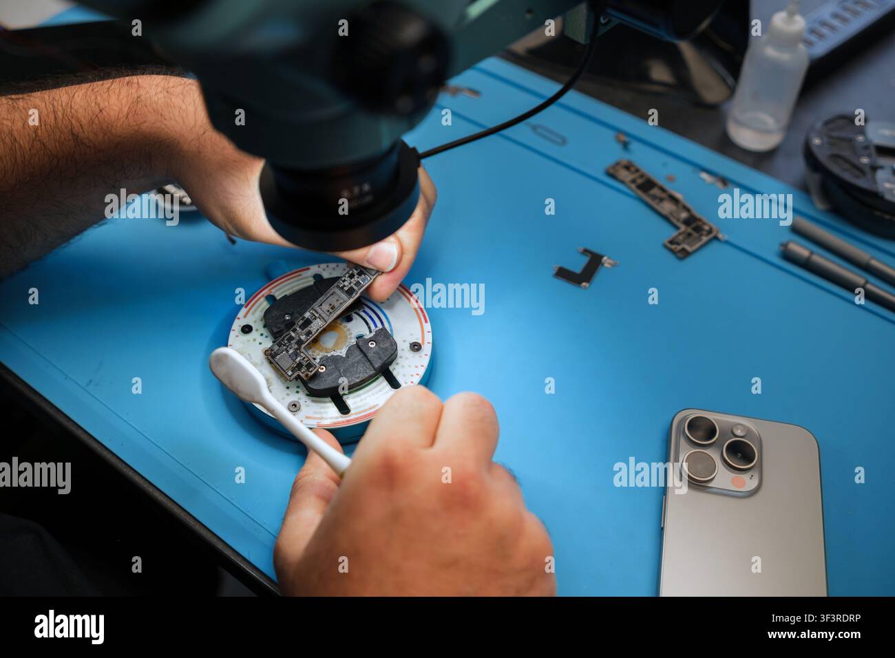 Technician repairs electronic device under microscope with tools and smartphone on blue work surface in a modern workshop setting Stock Photo