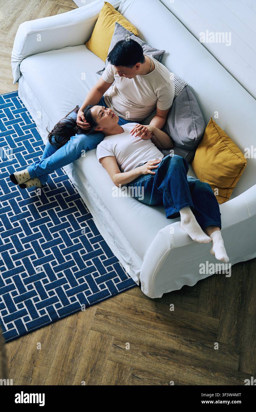 Overhead view of a couple relaxing together on a white sofa at home, sharing a quiet moment. Man rests beside woman as they hold hands in a cozy livin Stock Photo