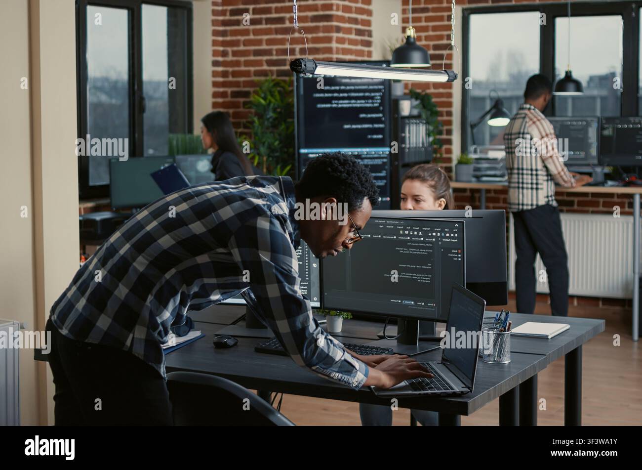 Senior developer codes on a laptop at a desk with multiple screens parsing software, compiling programming language and reviewing algorithm results. Stock Photo